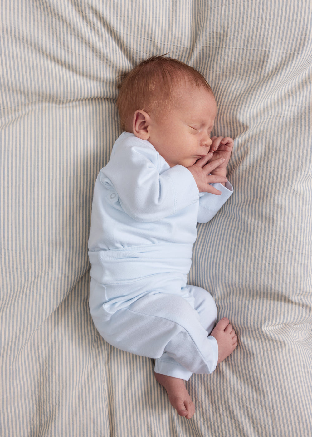 Babysengetøj med blå/hvide striber under sovende baby i hvid heldragt