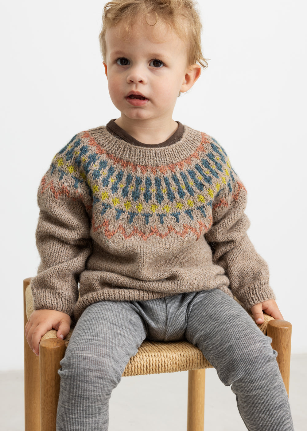 Dreng iført beige uldstrikbluse med mønster sidder på stol.