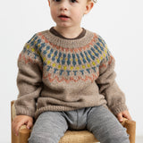 Dreng iført beige uldstrikbluse med mønster sidder på stol.