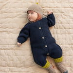 Baby ligger på tæppe iført beige uldhue og mørk holdragt.