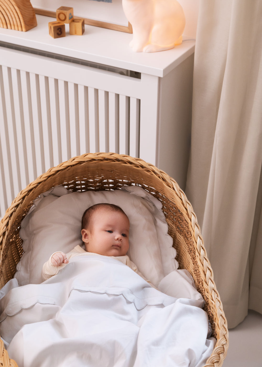 Babysengetøj i hvidt med skalkant; baby ligger i kurv med dyne og pude.