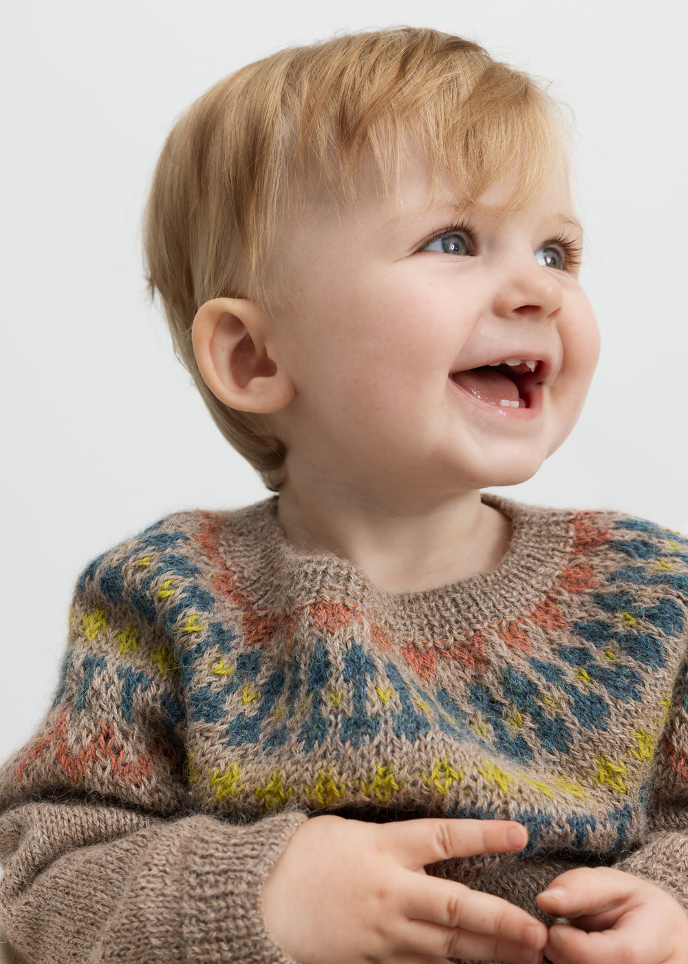 Baby smiler iført beige uldstrikbluse med farvet bærestykke