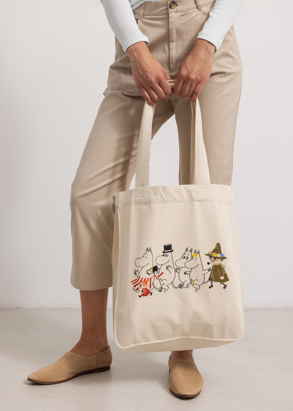 Creme tote-taske med Moomin-figurer på forsiden, vist i hænder og ved ben