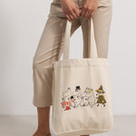 Creme tote-taske med Moomin-figurer på forsiden, vist i hænder og ved ben