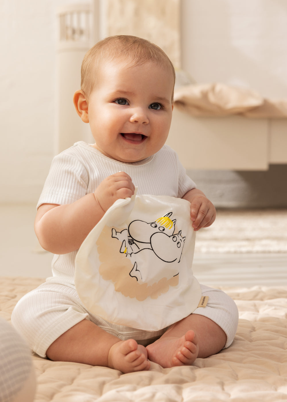 Hvidt knitrelegetøj med Mummi-print, holdt af baby på tæppe
