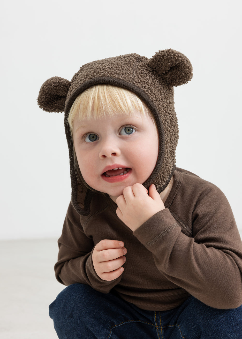 Barn iført brun elefanthue med ører i teddyfleece, sidder og ser op.