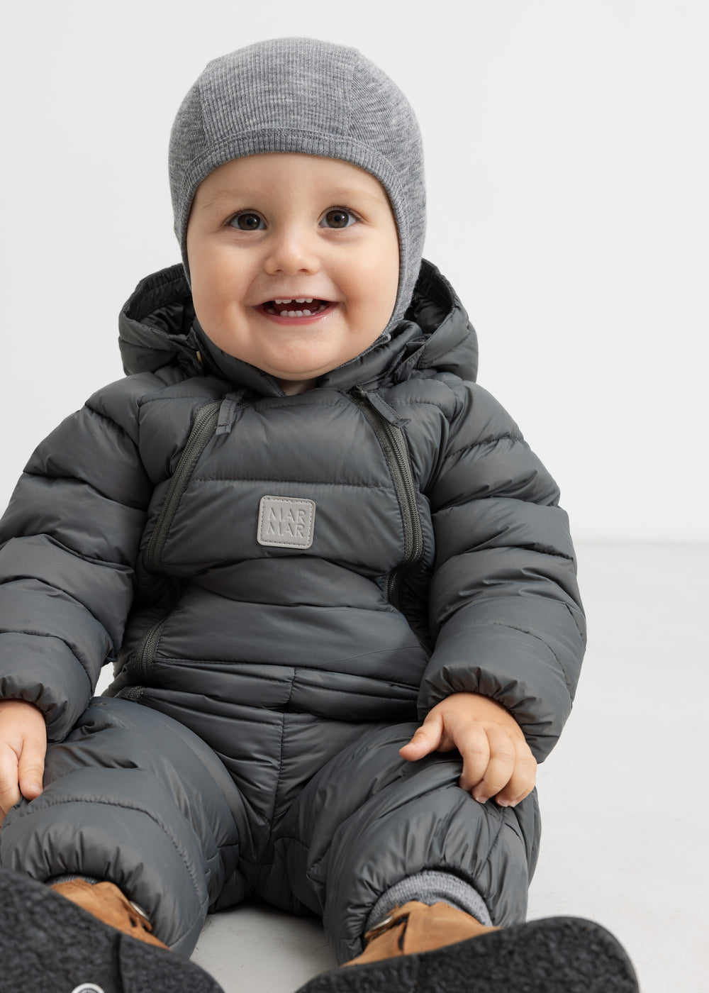 Smilende baby i mørkegrå dundragt sidder på gulv.
