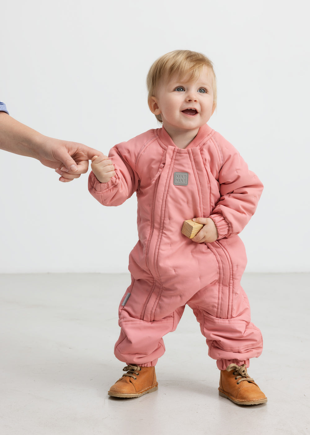 Baby iført lyserød termodragt, holder en voksen i hånden og smiler.