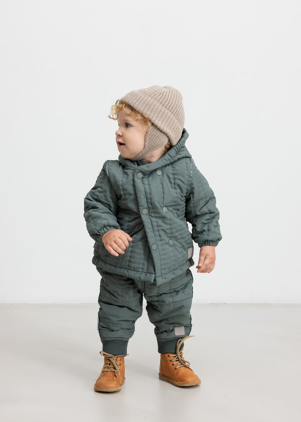 Baby iført støvet grøn termojakke og bukser, står og kigger til siden.