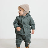 Baby iført støvet grønt termotøj og beige hue, står og kigger til siden.