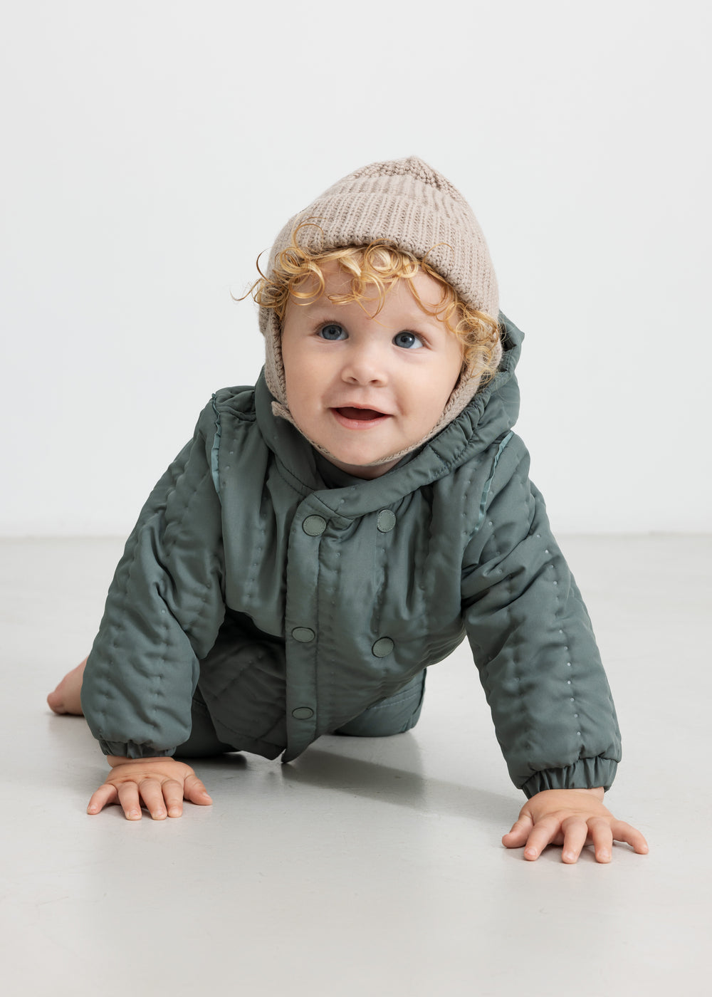 Baby iført beige strikhue og mørkegrøn heldragt, kravler mod kameraet.