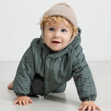 Baby iført beige strikhue og mørkegrøn heldragt, kravler mod kameraet.