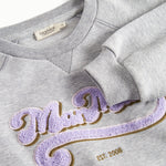 Nærbillede af lila broderet logo på grå sweatshirt til børn