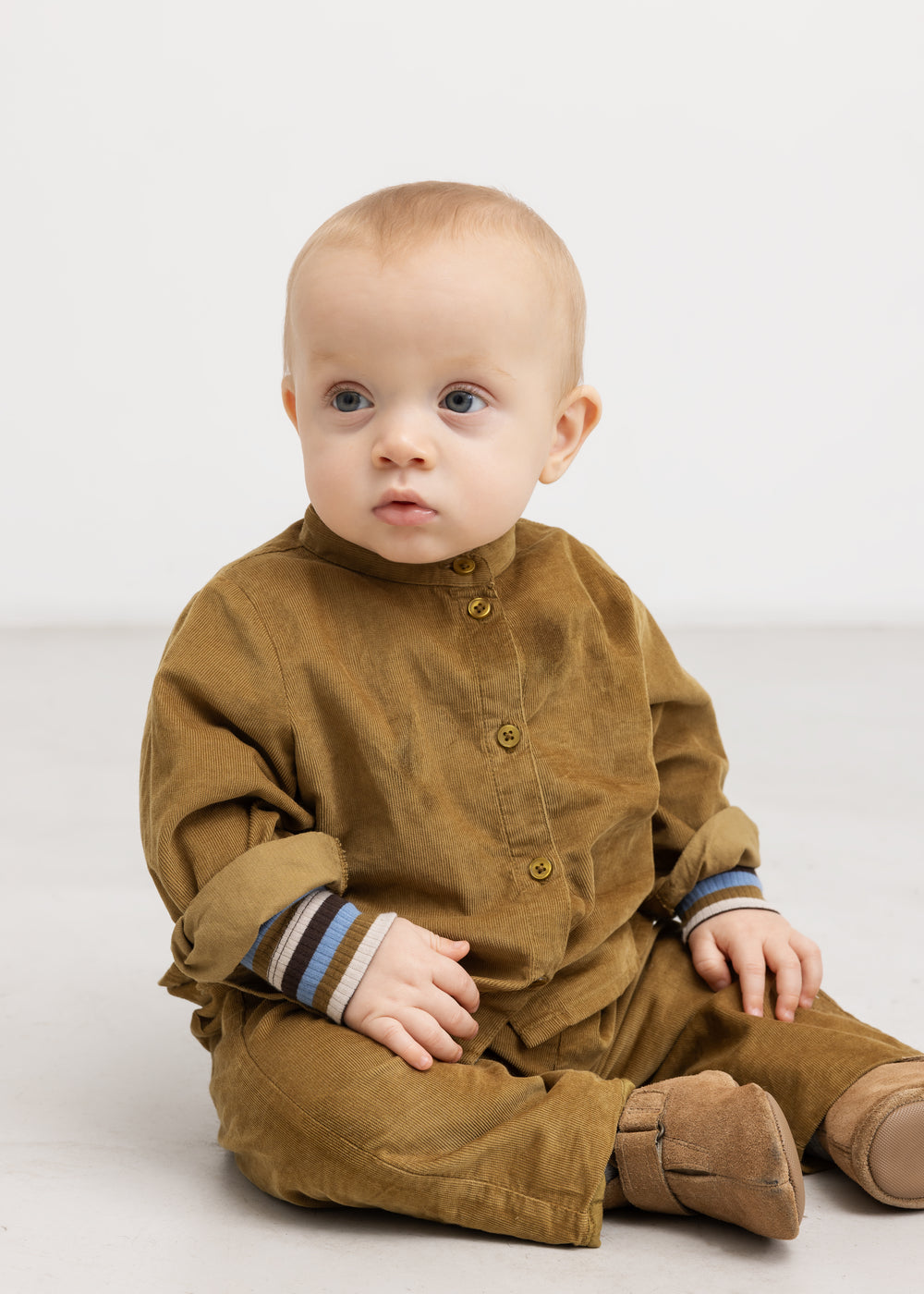 Baby iført langærmet bodystocking med stribede manchetter under brun heldragt/kjole-stil.