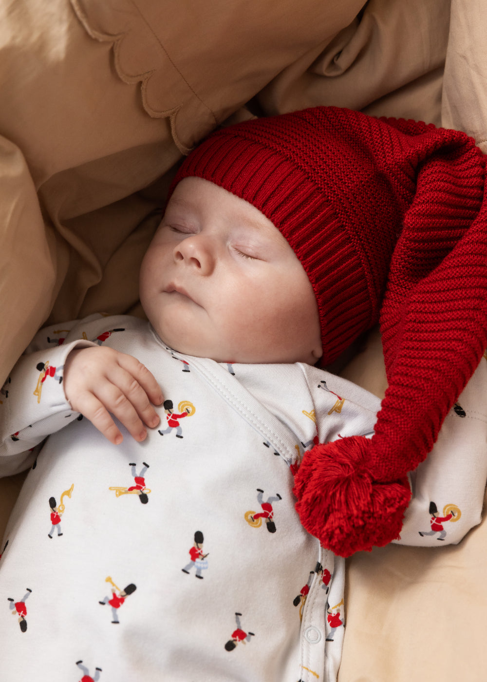 Baby i hvid heldragt med musiker-mønster og rød hue med pompon.