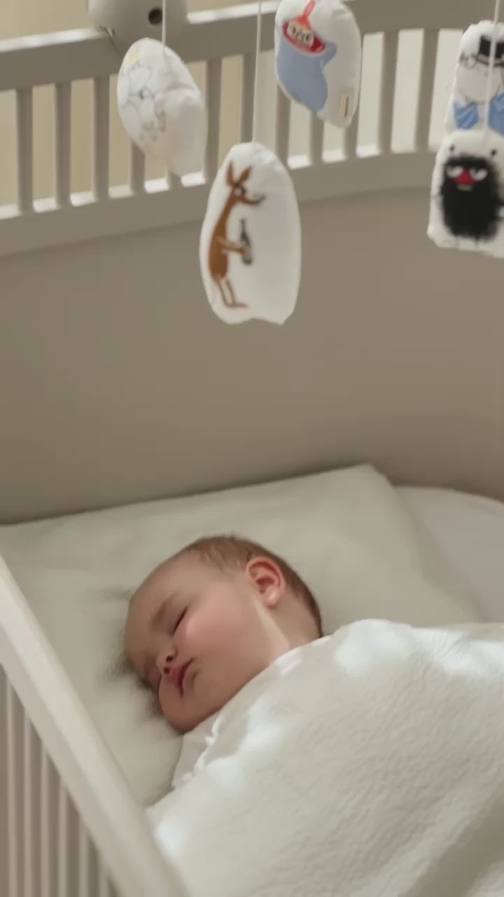 Baby sover i tremmeseng under hvidt tæppe med Moomin uro over